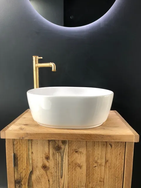 White round ceramic countertop basin with a gold mixer tap, mounted on a rustic wooden vanity unit in a contemporary bathroom.